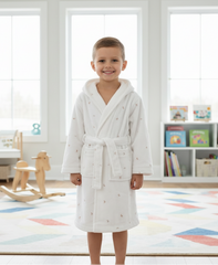 A child wearing a white robe with a hood in a room with a colorful rug and shelves.
