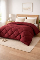 Red comforter on a bed in a bedroom with light wood flooring and a lamp.