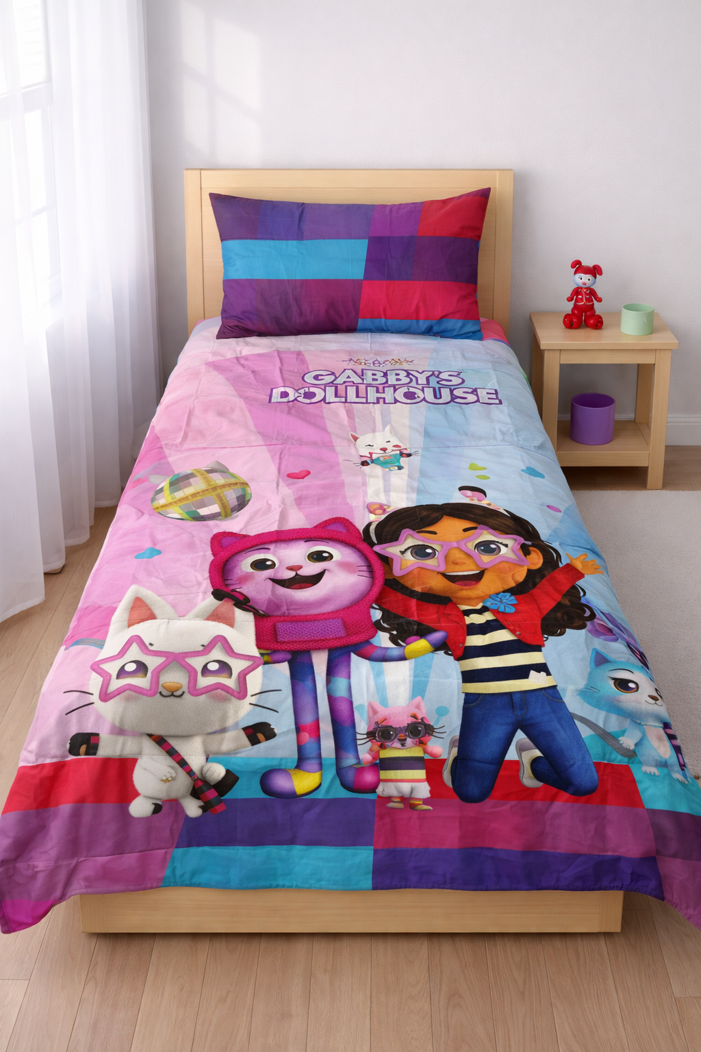 Children's bed with colorful bedding featuring cartoon characters, on a wooden floor with a white curtain.