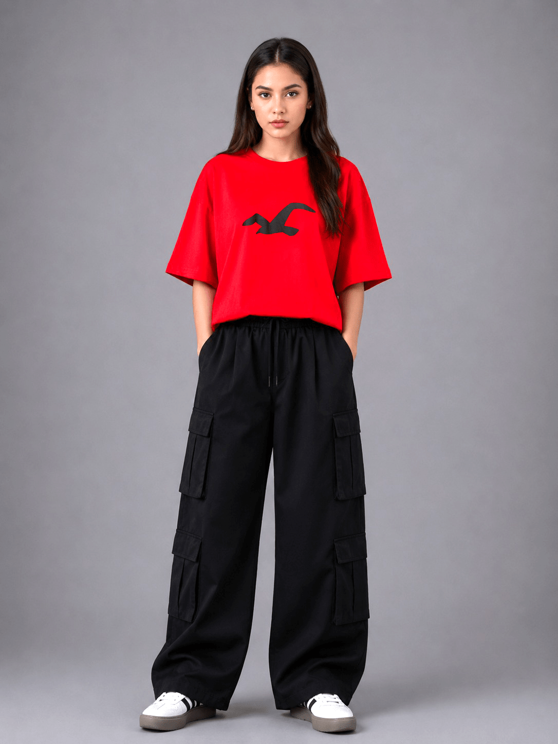 A model stands against a gray studio background wearing a bright red short-sleeve t-shirt with a black bird logo on the chest, paired with black wide-leg cargo pants and white sneakers.