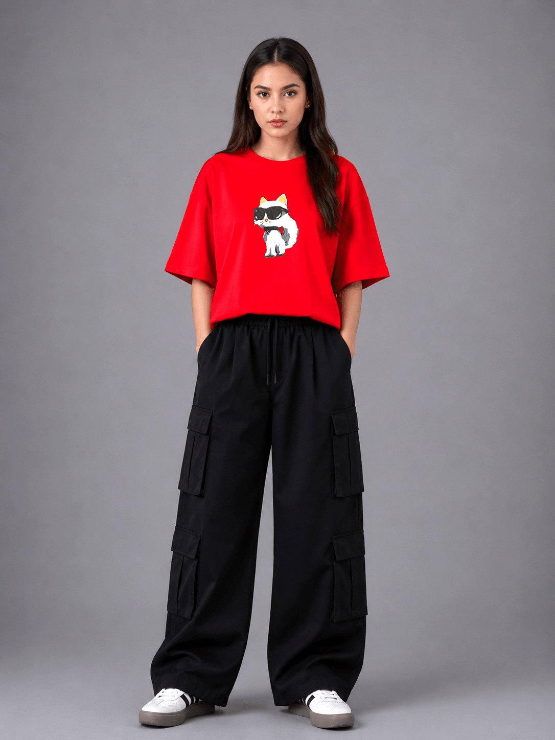 A woman stands against a gray studio background wearing a bright red short-sleeve T-shirt with a white and black cat graphic on the chest, paired with loose black cargo pants and white sneakers.