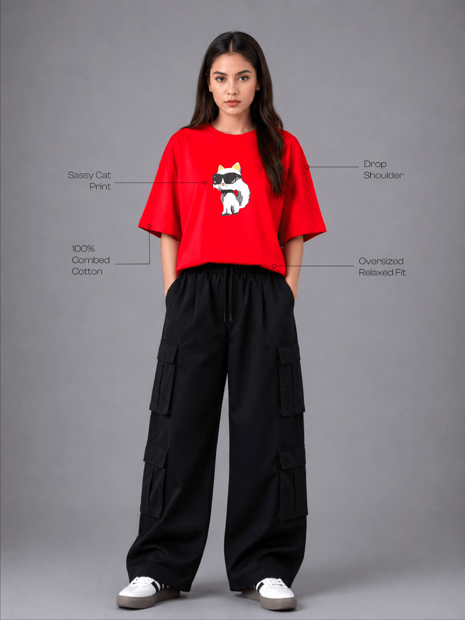 A model stands against a gray studio background wearing a bright red short-sleeve T-shirt with a white cat graphic and black wide-leg cargo pants. Text callouts point to features of the shirt, including 'Sassy Cat Print,' 'Drop Shoulder,' '100% Combed Cotton,' and 'Oversized Relaxed Fit.